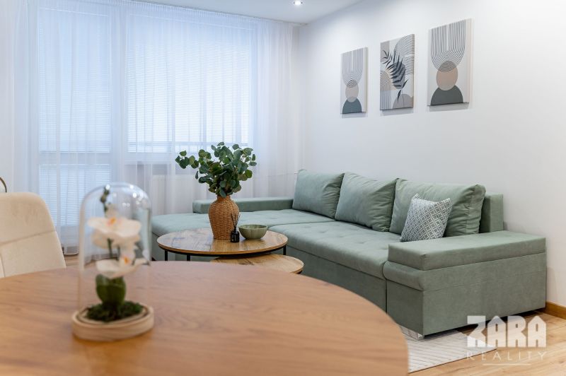 A living room in a two-room apartment with a green sofa, flowers, and paintings on the wall.