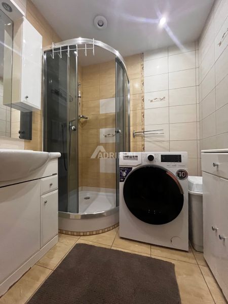A bathroom in a 2-room apartment with a washing machine, shower, and light tiles.