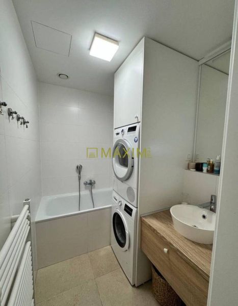 A bathroom with a washing machine, dryer, and bathtub in a 3-room apartment, light tiles, and wooden decor.