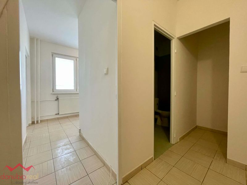 A hallway in a one-room apartment with tiled flooring, white walls, and a large window.