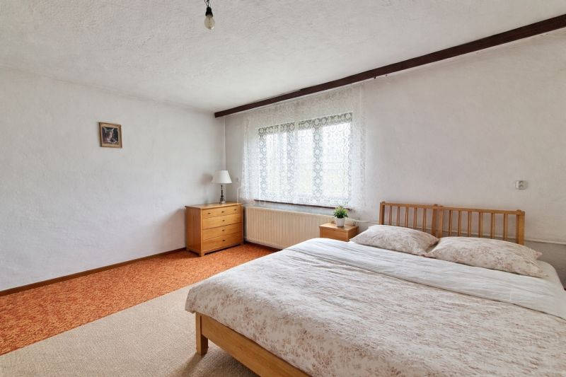 Bedroom in a family house with a wooden bed and dresser, white walls, small window.