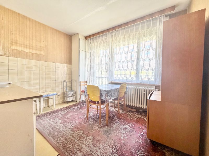 A kitchen in a 2-room apartment with a table, a carpet, and a large window with lace curtains.