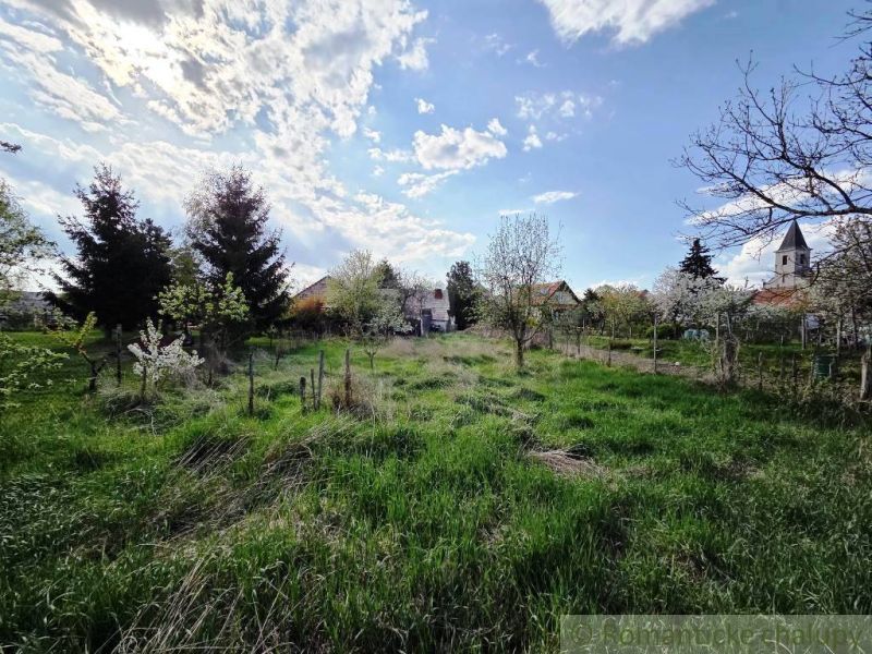 A blooming garden in Šahy surrounds cottages and a church under a blue sky.