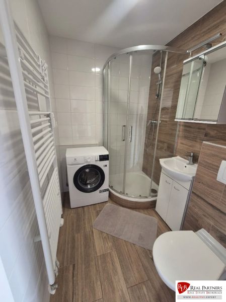 Bathroom of a studio apartment with a shower, washing machine, and floor with a wood decor.