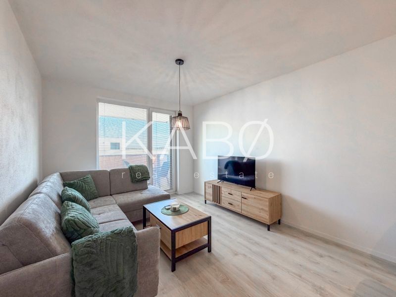 Living room in a two-room apartment with a large couch, television, and wooden-patterned flooring.