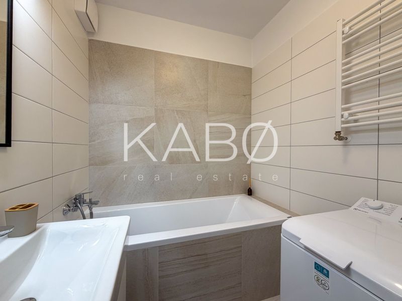 A bathroom in a 2-room apartment with a bathtub, sink, and washing machine, tiled with light tiles.