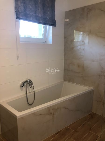 A bathroom in a family house with a bathtub, tiles, and a window. The floor has a wooden decor.