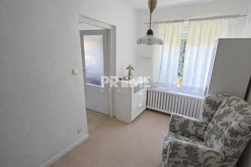 A room in a family house with an armchair, drawers, and a large window with white curtains.