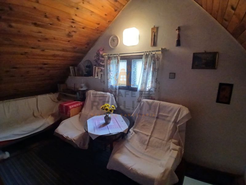A room with a slanted ceiling, a sofa with upholstery, and a chair by a table with a vase.