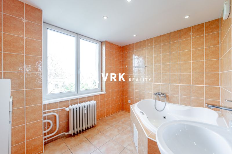 A bathroom in a 2-room apartment with a corner bathtub, beige tiles, and a large window.