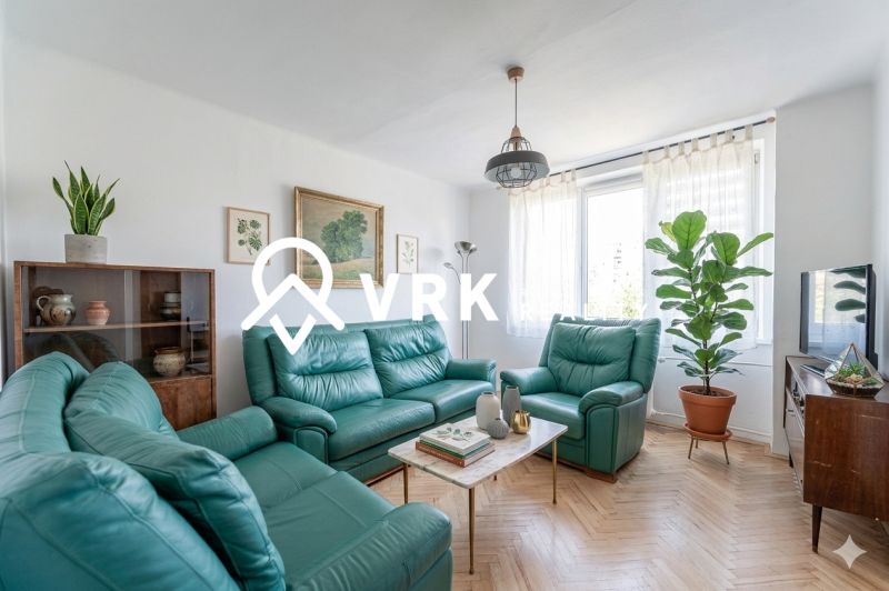 Living room in a two-room apartment with sofas, plants, and a wooden decor floor.