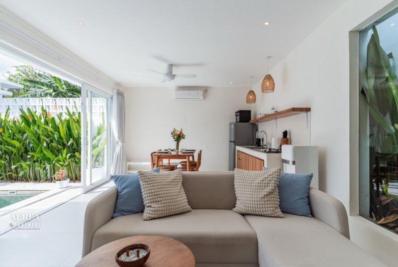 Interior of a villa with a white sofa, kitchen, and view of the pool and greenery.