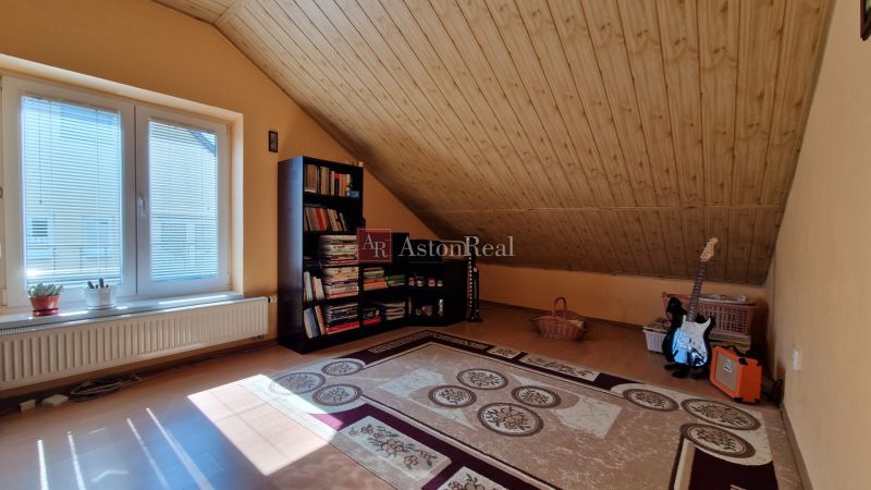 Attic room in a family house with wooden ceiling decor, carpet, and a bookshelf full of books.