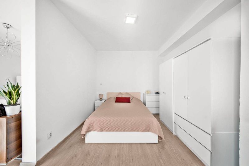 Bedroom in a two-room apartment with a wooden decor floor, white furniture, and a plant.