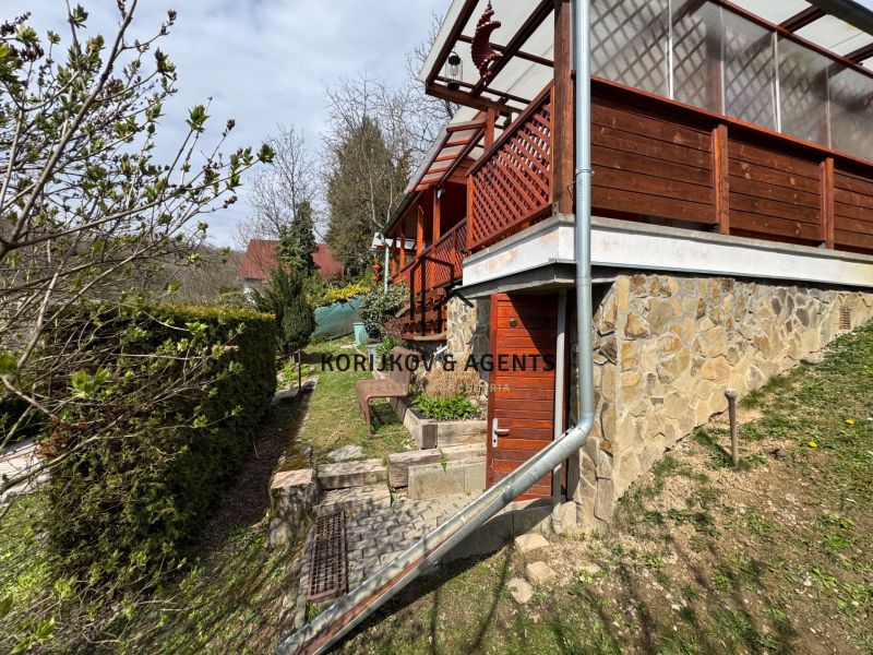 A cottage in Svinica with a stone facade and a wooden terrace, surrounded by greenery.