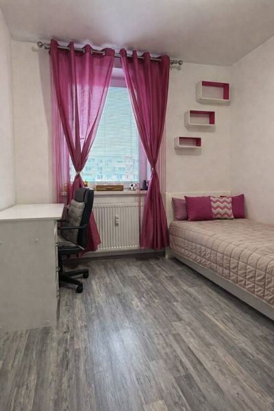 A room in a 4-room apartment with pink curtains, a bed, a table, and a wooden decor floor.