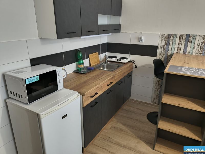 Kitchen area of a studio apartment with appliances and flooring with a wood decor.