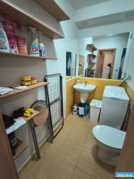 Studio apartment bathroom with shelves, an appliance, and a toilet, floor with wood decor.