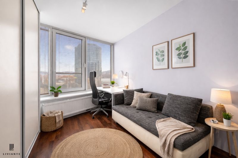 A room with a sofa, a desk, a wooden patterned floor, and large windows.