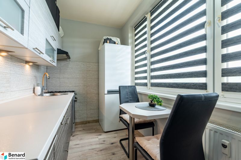 A kitchen in a 2-room apartment with a table, chairs, a kitchen unit, and a refrigerator, with a wooden decor floor.
