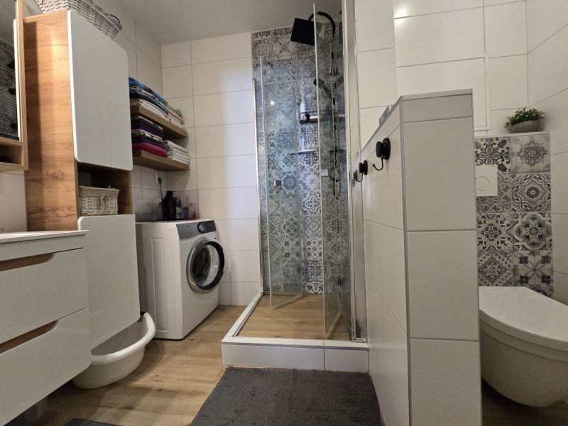 A bathroom in a 2-room apartment with a washing machine, shower, and wooden decor flooring.
