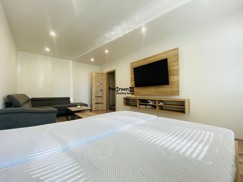 Living room in a two-room apartment with a dark sofa, TV, and wooden decor flooring.
