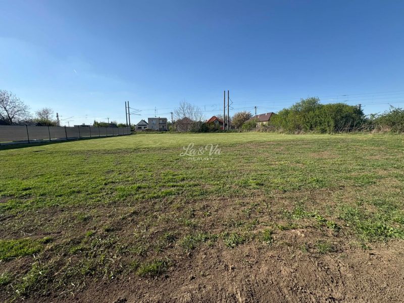 A flat plot of land in Pozba designated for residential use, with a view of neighboring houses.