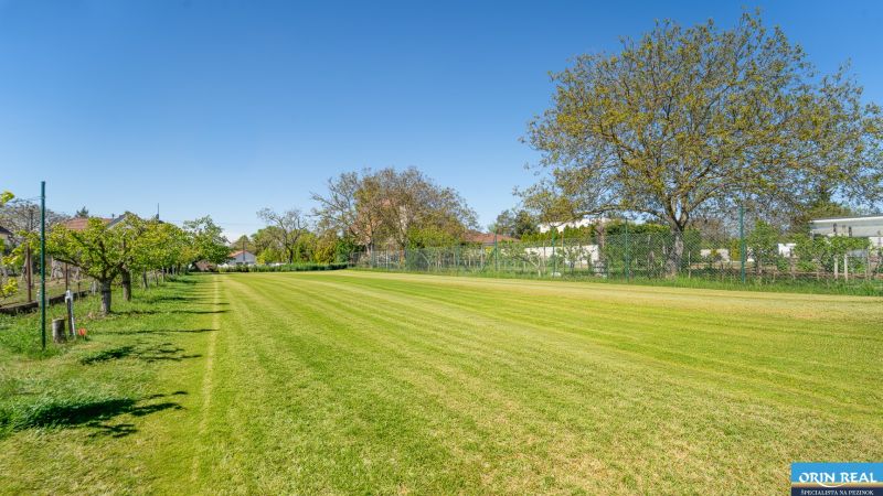 A grassy residential plot on Nová Street in Viničné, bordered by trees and a fence.