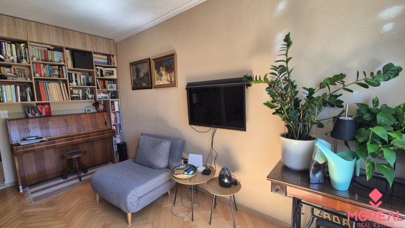 Living room of a two-room apartment with a wooden decor floor, a bookshelf, a piano, and plants.
