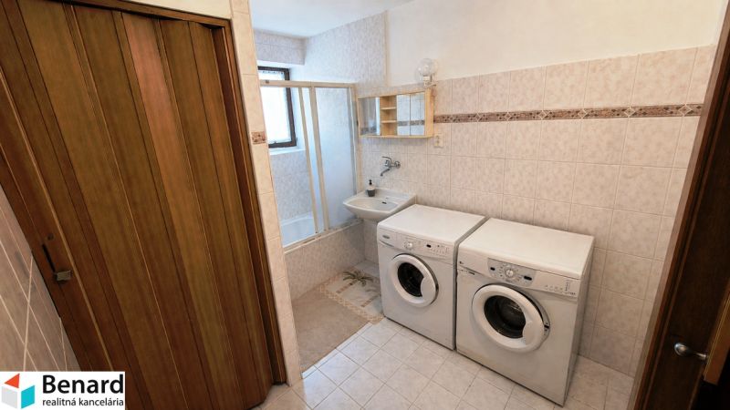 A bathroom in a family house with a washing machine, dryer, and bathtub on light tiles.