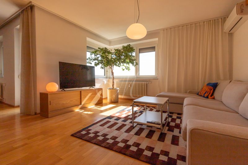 Living room in a 4-room apartment with a sofa, television, carpet, and wooden decor flooring.