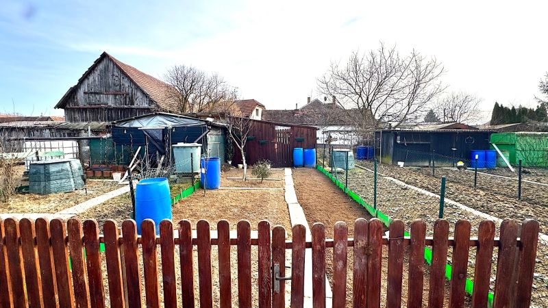 A garden in Hlboké with rural buildings and barrels between fences.