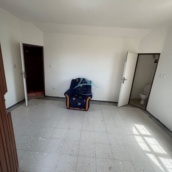 A room with a chair, white walls, tiles, and a door leading to the toilet in the cottage.