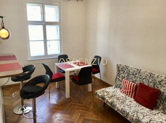 Living area in a two-room apartment with a wood-patterned floor and a dining table.