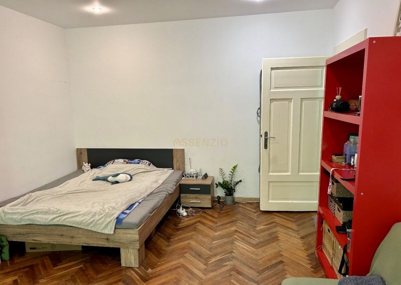 Bedroom in a 2-room apartment with a bed, red shelf, and wooden decor flooring.