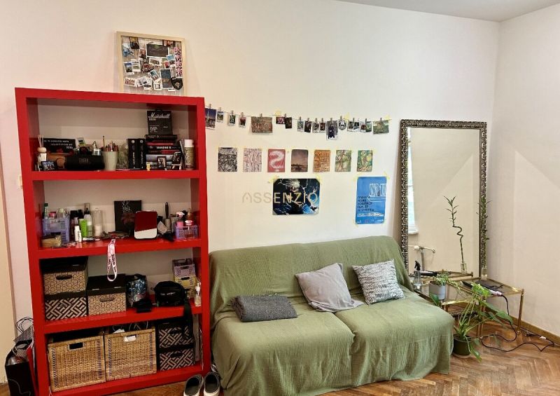 A living room in a two-room apartment with a green sofa, a red shelf, and a wooden decor floor.