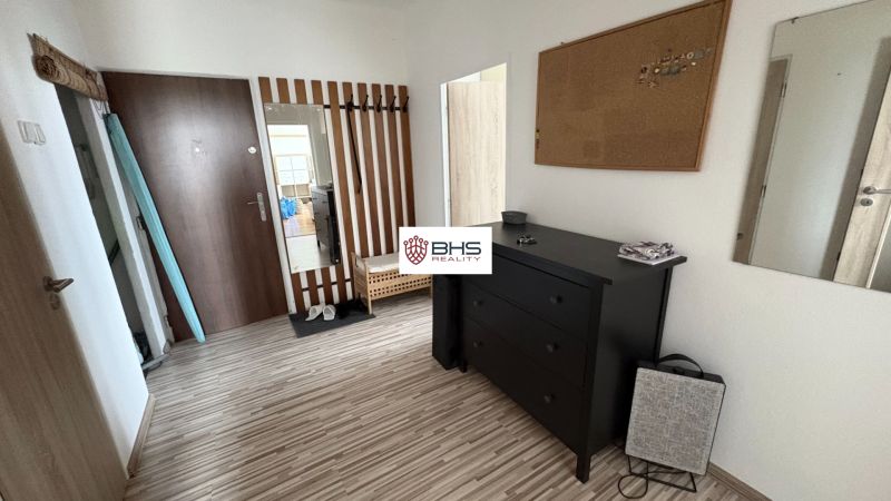 A hallway in a 2-room apartment with a wooden-patterned floor, a mirror, and a black dresser.