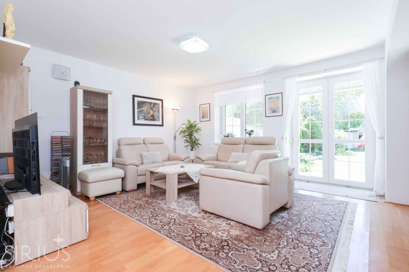 Living room of a family house with a light-colored sofa set, wood-patterned flooring, and large windows.