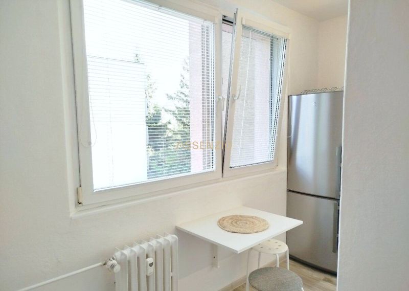 A kitchen in a studio apartment with a window, a refrigerator, and a small table for seating.
