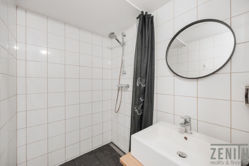 A bathroom with white tiles, a shower, and a sink in a two-room apartment.
