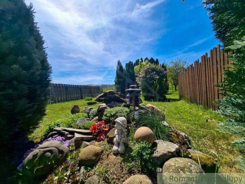 The garden of a family house in Mýtna with trees, a sculpture, and a wooden fence.