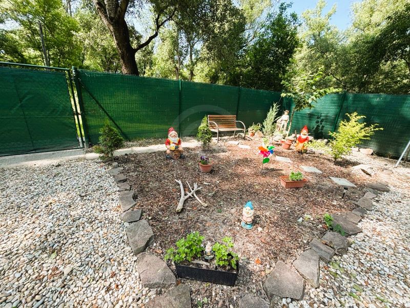 A garden with gnomes and a bench near a cottage in Gabčíkovo, surrounded by stones and a fence.