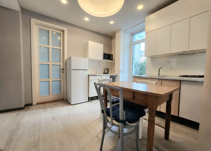 A kitchen in a 2-room apartment with a wooden decor floor, white cabinets, and a window.