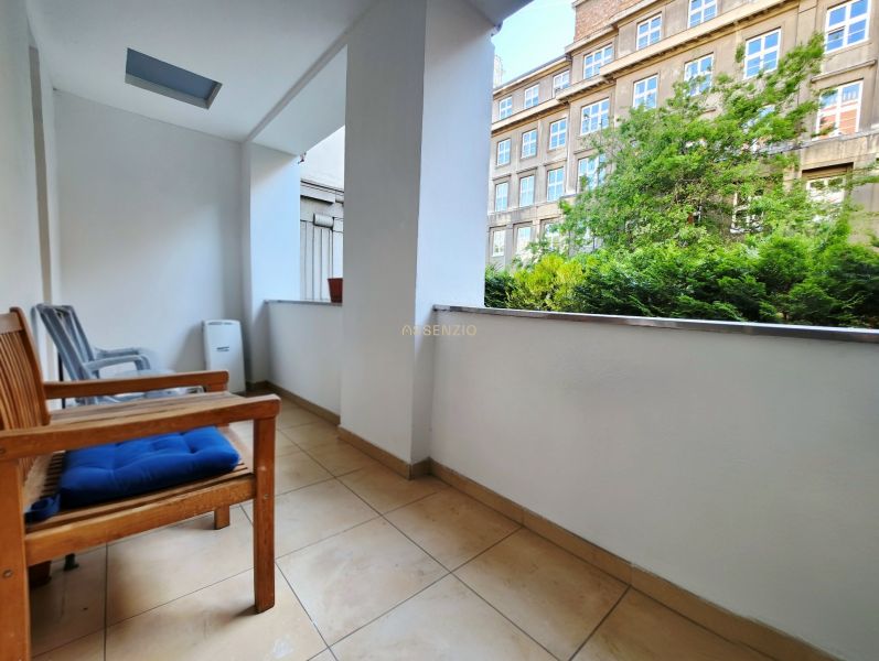 A balcony of a 2-room apartment with a view of greenery and a building, seating with chairs.