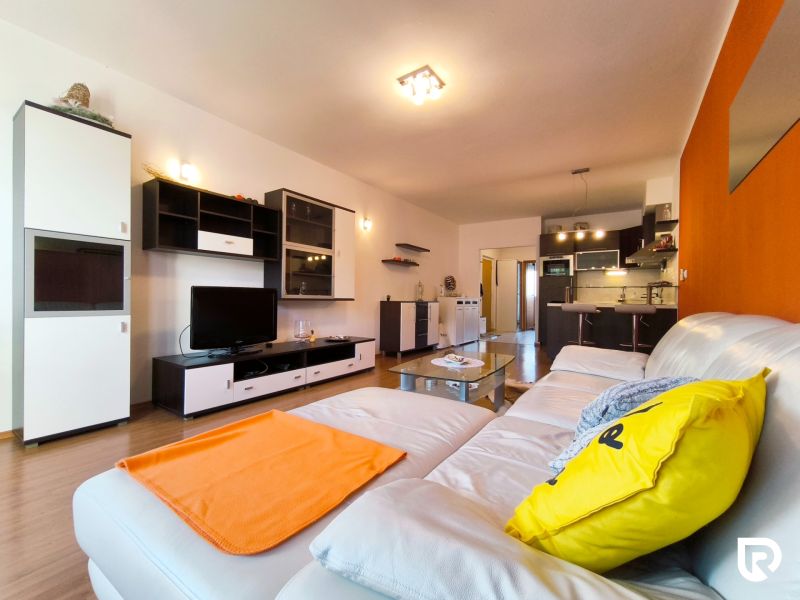 Living room in a 2-room apartment with a white sofa, orange accent, and a floor with a wood decor.