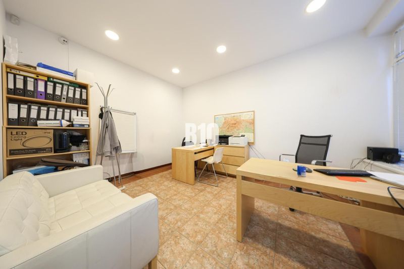 An office with wooden desks, a shelf with documents, a white chair, and a map on the wall.
