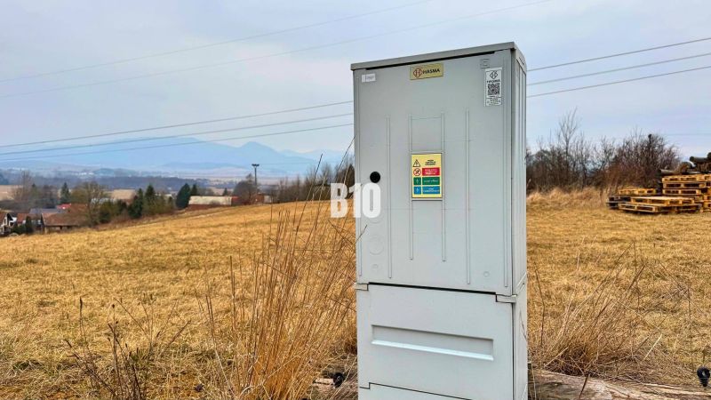 An electrical cabinet on a meadow, with hills and rural housing in the background.