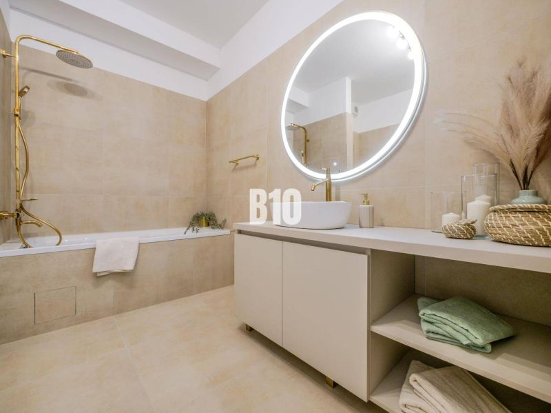 A bathroom in a 2-room apartment with a bathtub, a round mirror, and beige tiles.
