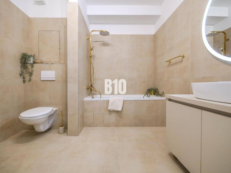 A bathroom in a 2-room apartment with beige tiles, a built-in bathtub, and a wall-mounted toilet.