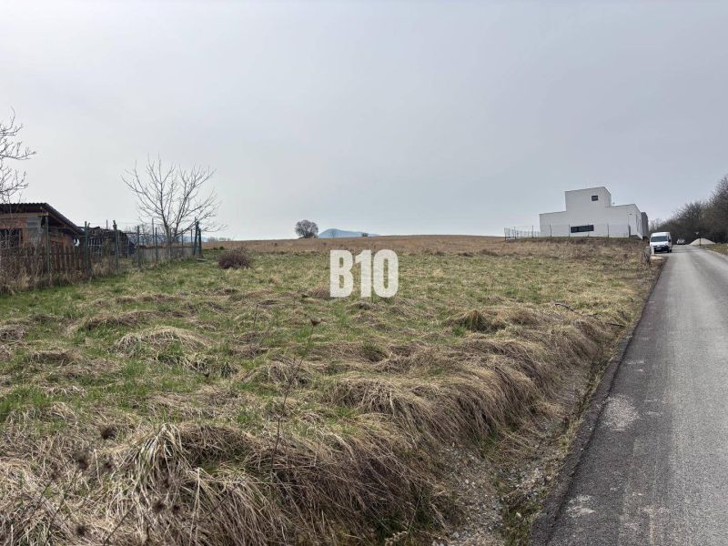 A plot for residential use in Rosina, next to the road, with a grassy surface and nearby buildings.